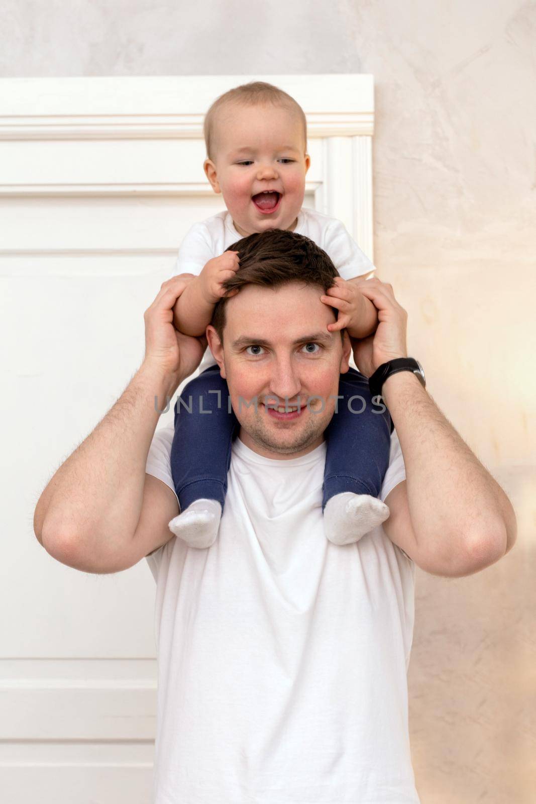 Bright guy in casual wear with excited son on shoulders standing at home looking at camera