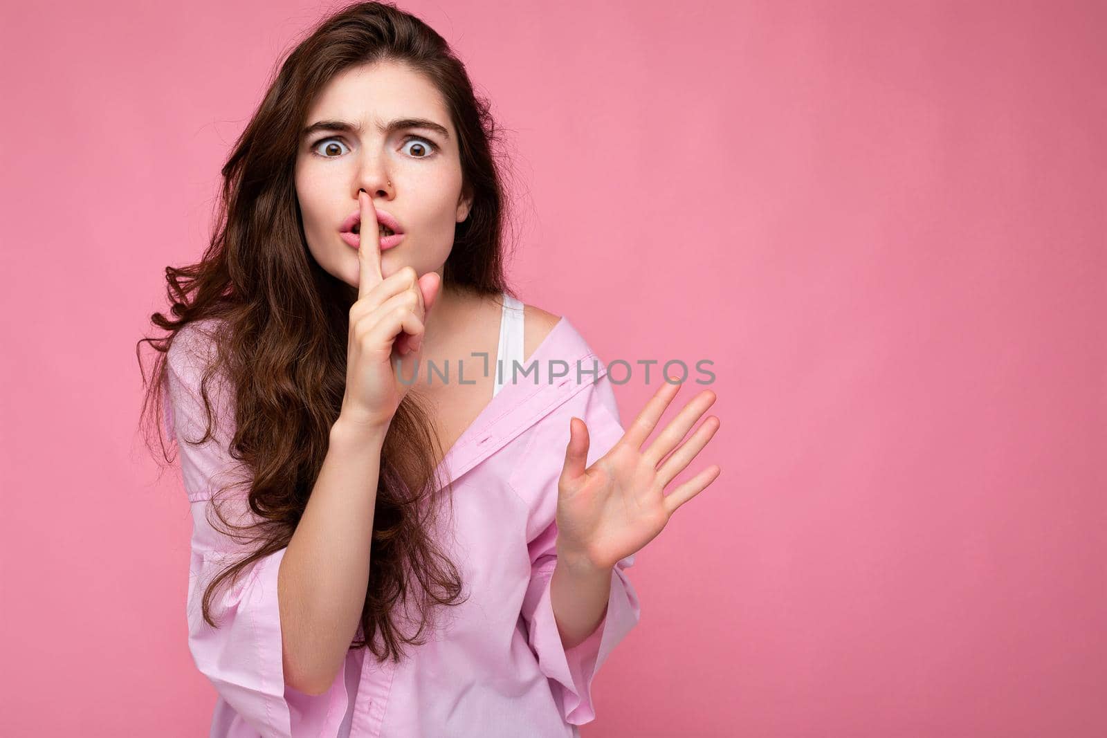 Photo of young emotional nervous beautiful wavy-haired brunette woman with sincere emotions wearing casual pink shirt isolated over pink background with copy space and showing silence gesture with finger near lips. Shhh sign.