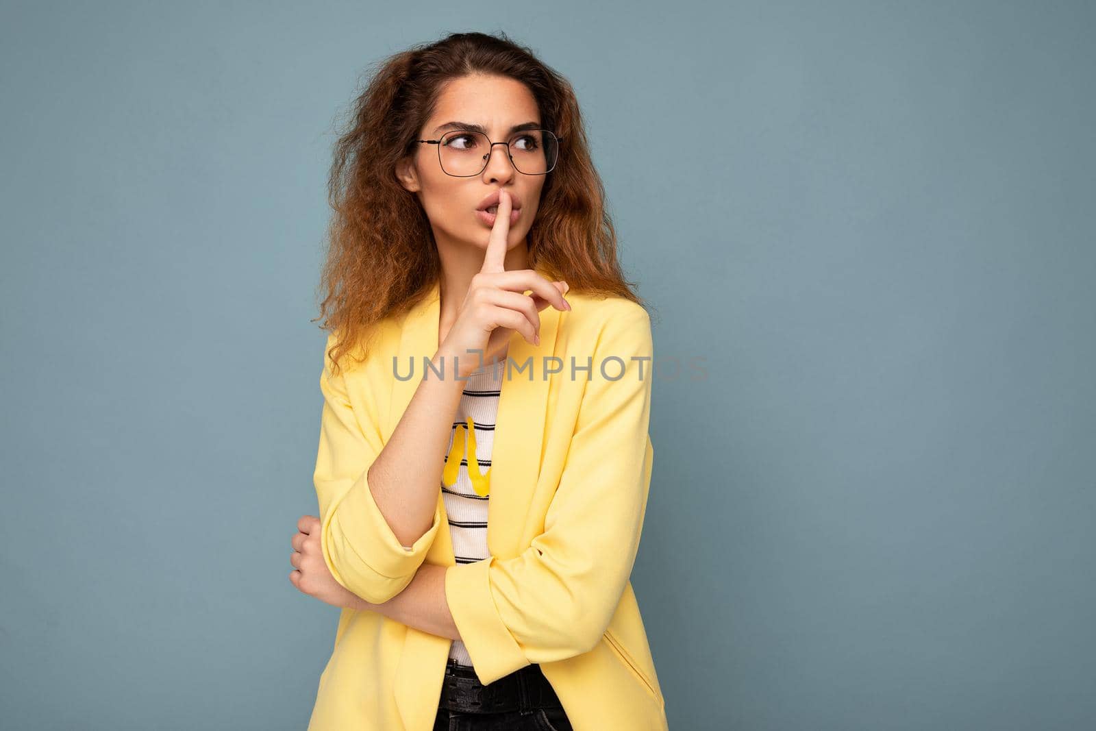 Photo of young beautiful brunette curly woman with sincere emotions wearing casual yellow jacket and optical glasses isolated on blue background with copy space and showing shhh gesture.