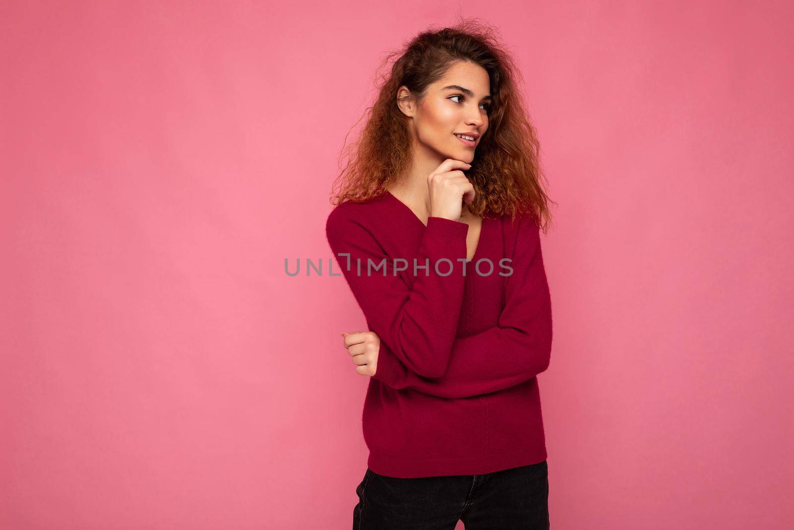 Photo of young european thoughtful fascinating brunette curly woman with sincere emotions wearing trendy pink sweater isolated over pink background with free space.