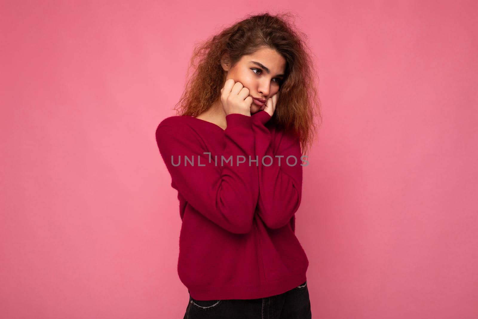 Portrait of young emotional offended touchy beautiful brunette curly woman with sincere emotions wearing trendy pink sweater isolated on pink background with copy space and taking offence.