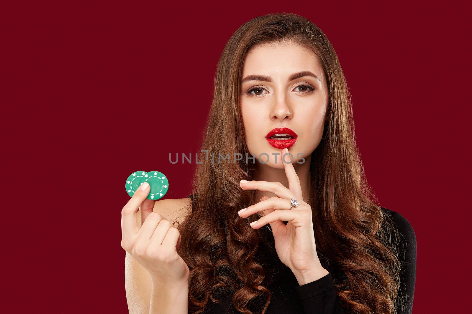 Pretty long hair woman in black dress holding chips for gambling in casino. Studio shot on red background. Poker. Two green Chips in Hand