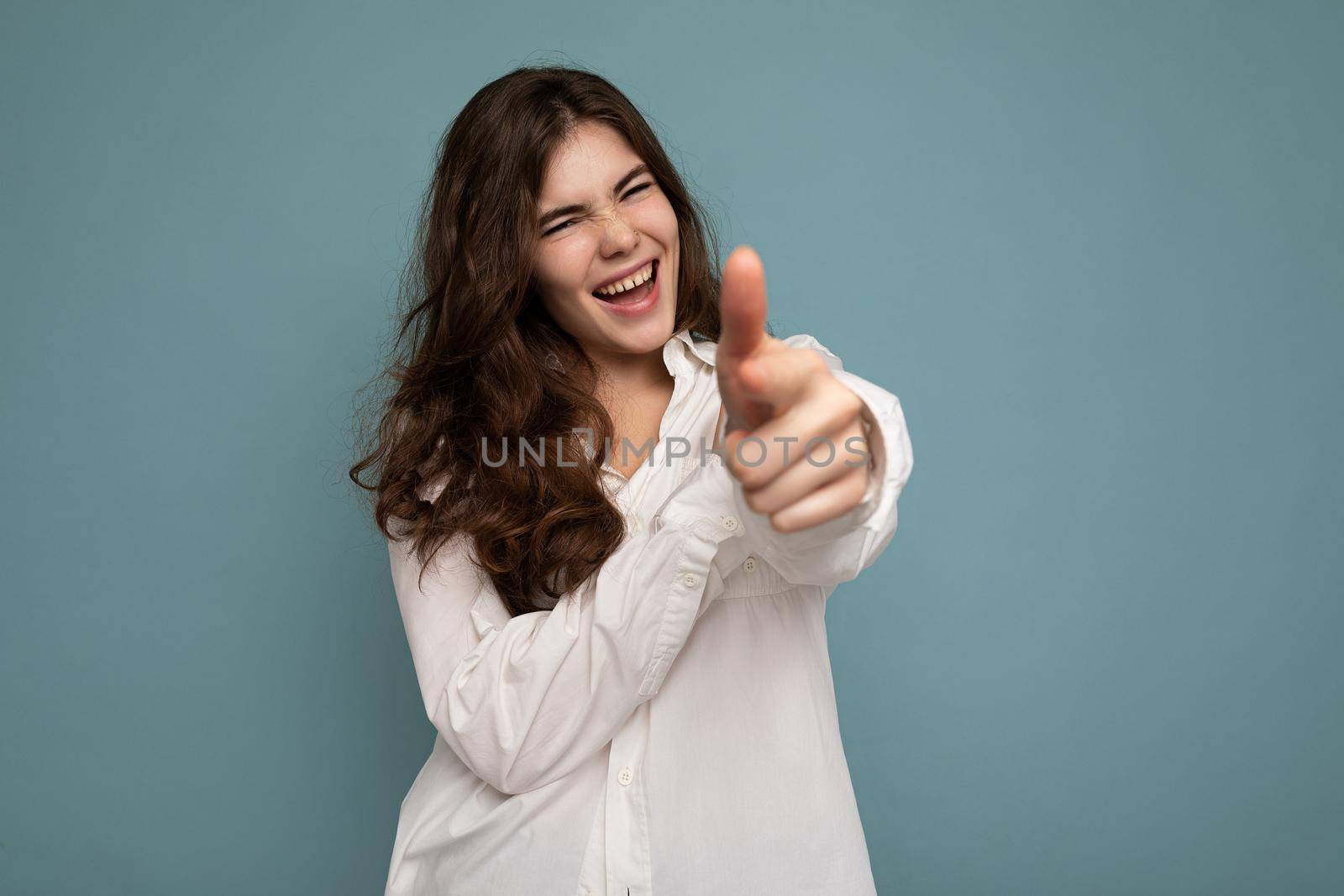 Portrait of young positive happy joyful attractive curly brunette woman with sincere emotions wearing casual white shirt isolated on blue background with empty space and pointing at camera.