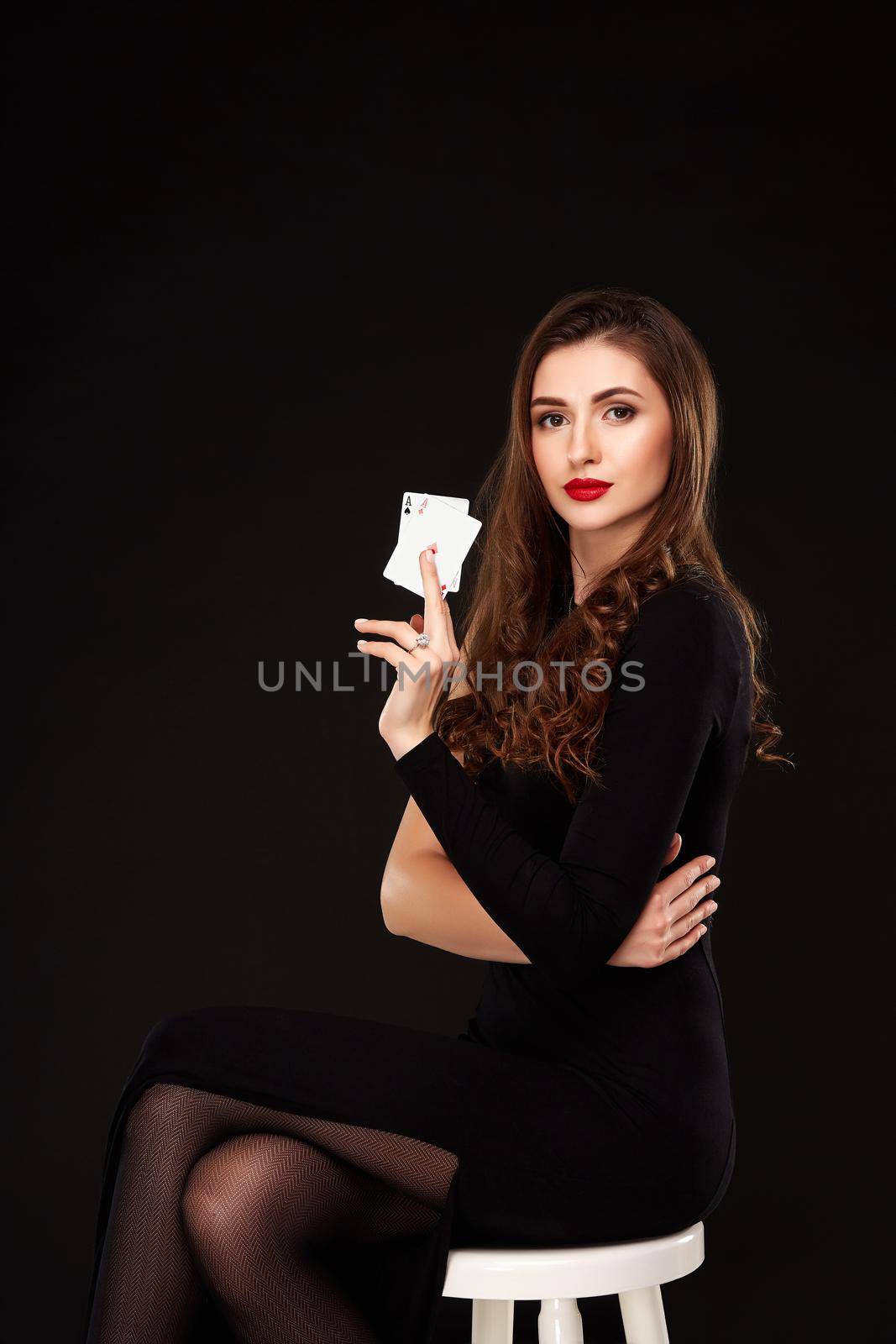 Sexy curly hair brunette in black dress posing with two aces cards in her hands, poker concept on black background. winning combination. sits on a chair