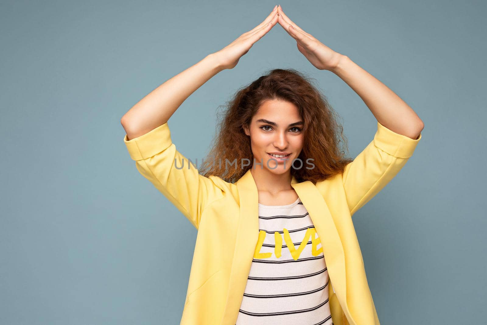 Photo shot of young positive happy beautiful brunette curly woman with sincere emotions wearing trendy yellow jacket isolated on blue background with copy space and showing house gesture with hands. Home concept.