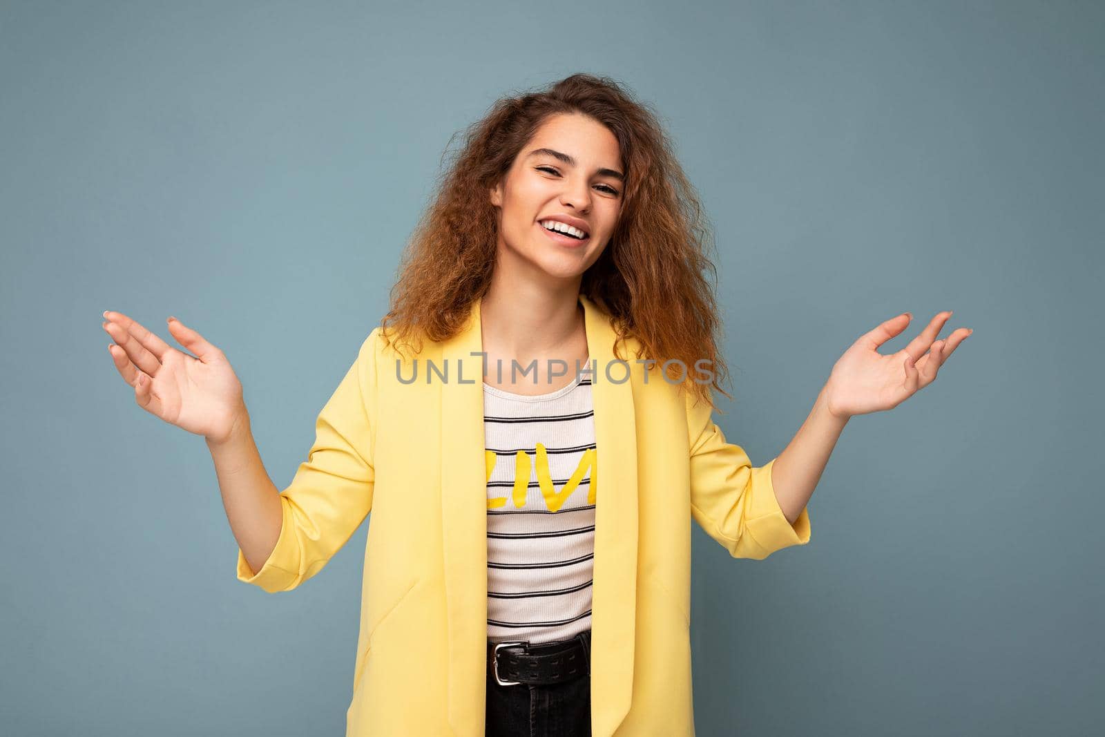 Photo shot of young smiling positive happy attractive brunette curly woman with sincere emotions wearing trendy yellow jacket isolated on blue background with empty space and having doubts.