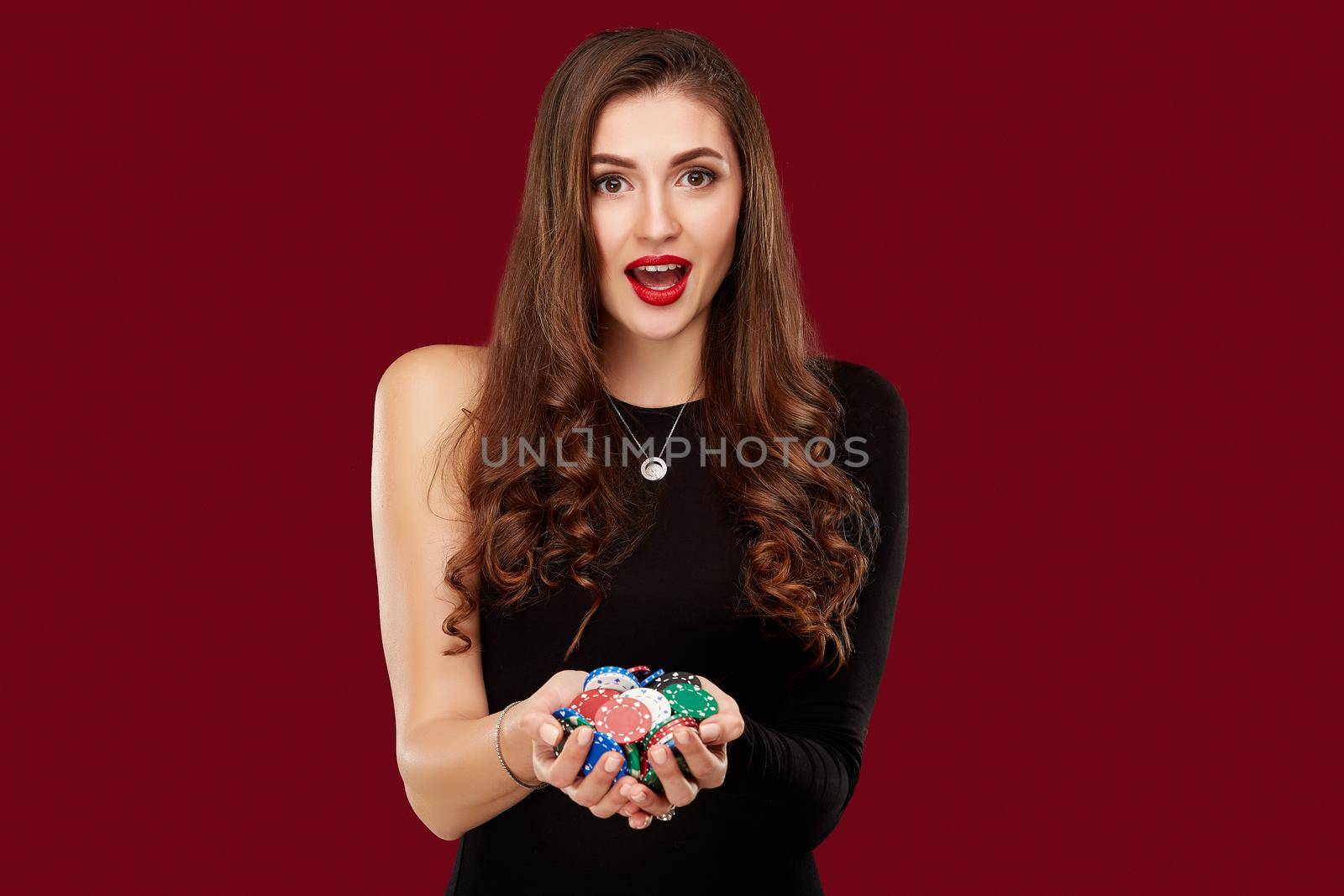 Casino, gambling, poker, people and entertainment concept - woman poker player in black dress with chips in hands on red background. Studio shot