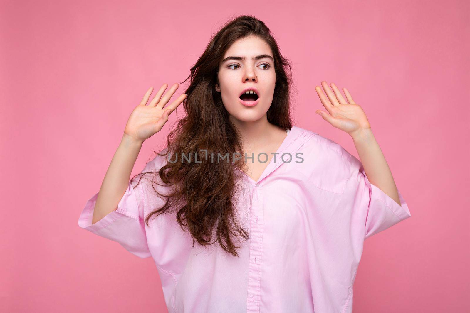 Portrait of young emotional shocked surprised winsome nice curly brunette woman with sincere emotions wearing casual pink shirt isolated over pink background with empty space. I don't understand