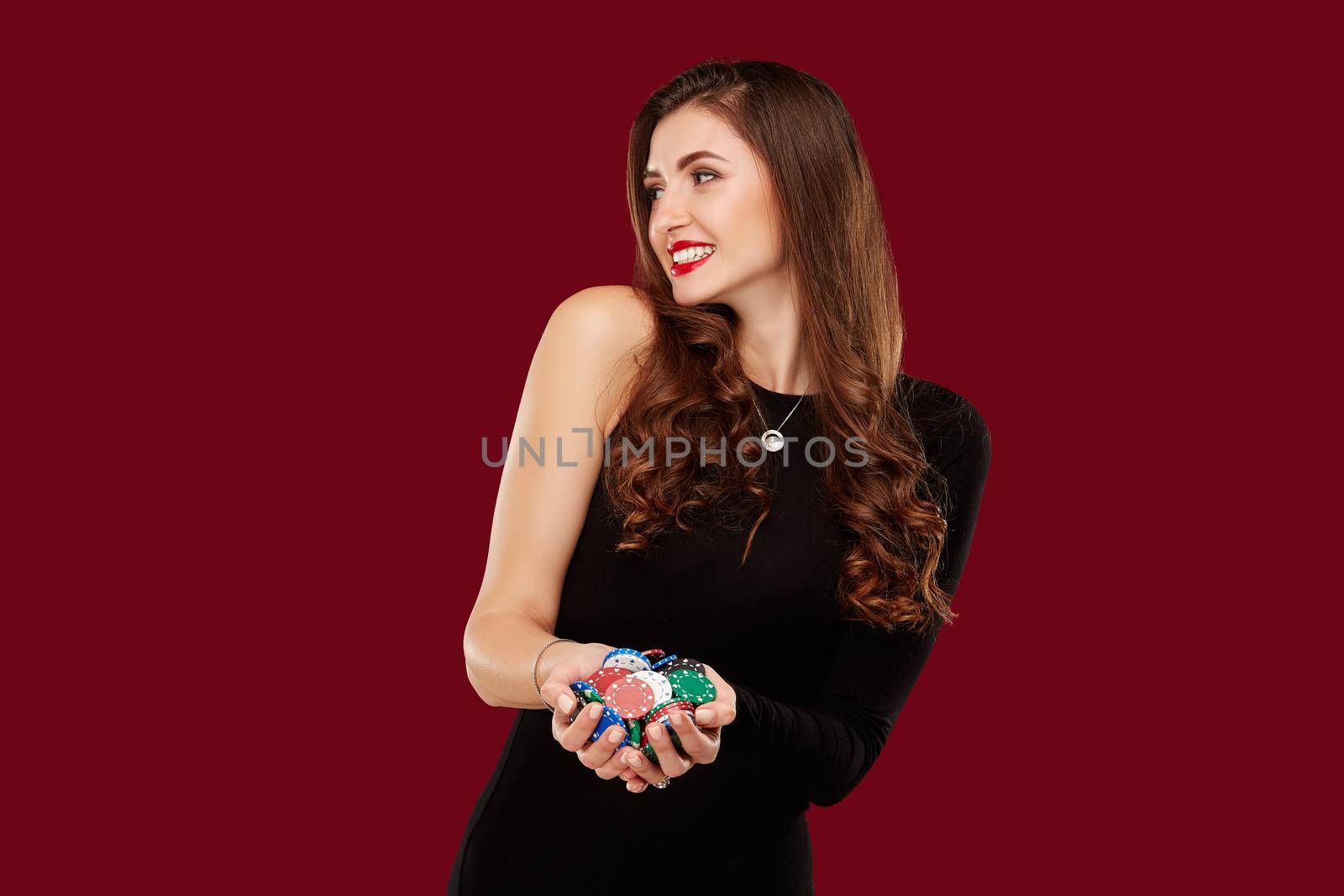 Casino, gambling, poker, people and entertainment concept - woman poker player in black dress with chips in hands on red background. Studio shot