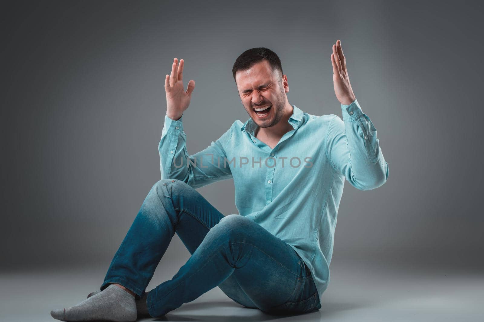 Handsome young man sitting on a floor with raised hands gesturing happiness on gray background. A man in jeans and a blue shirt