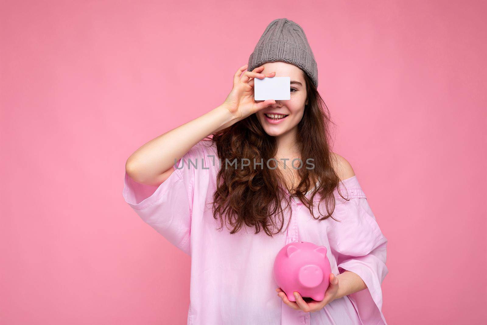 Portrait of beautiful positive cheerful cute smiling young brunette woman in stylish shirt isolated on pink background with copy space and holding pink pig moneybox and credit card for mockup.