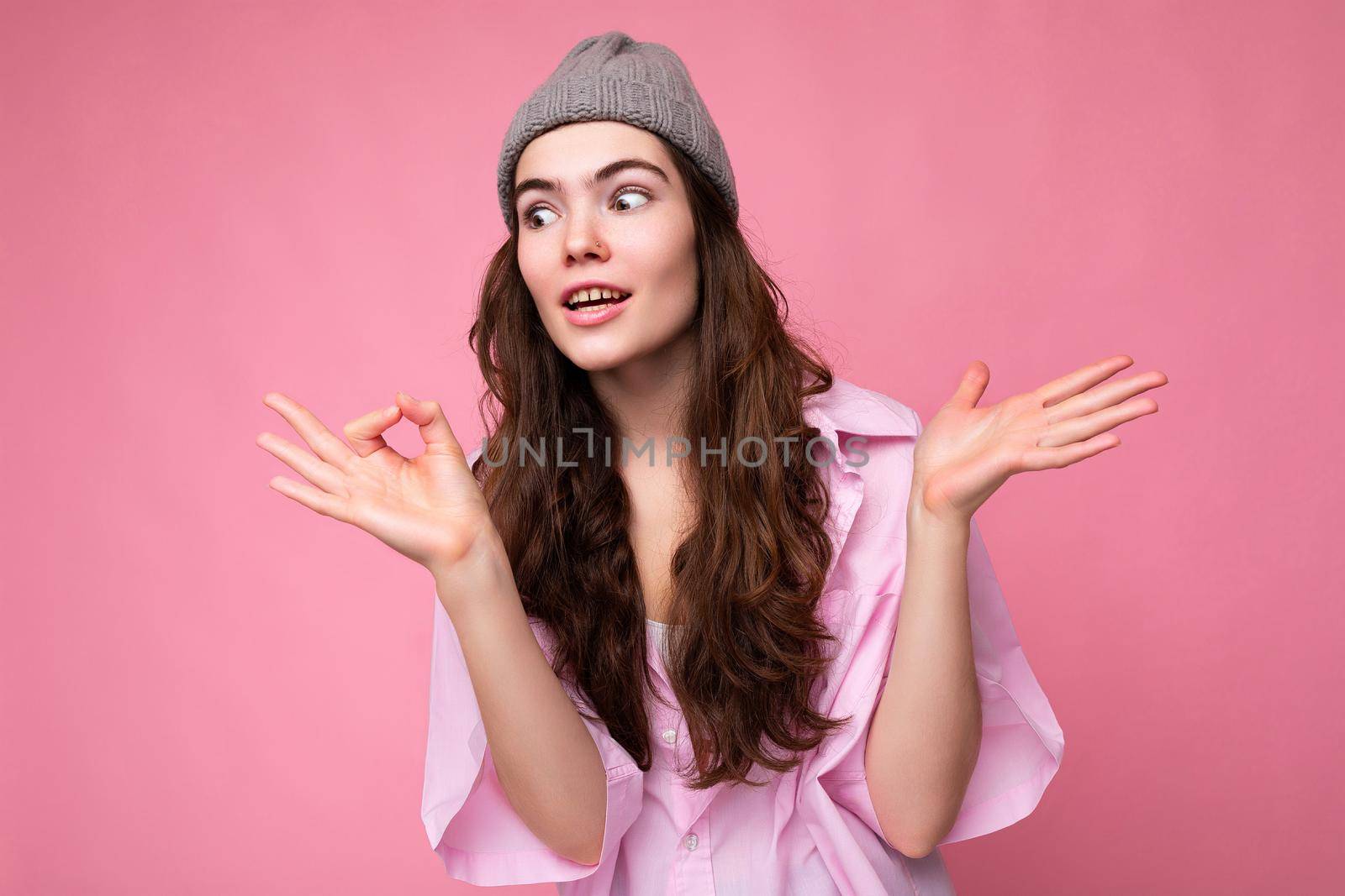 Beautiful strange funny young curly brunette woman wearing pink shirt and grey hat isolated on pink background with copy space and don't understanding.