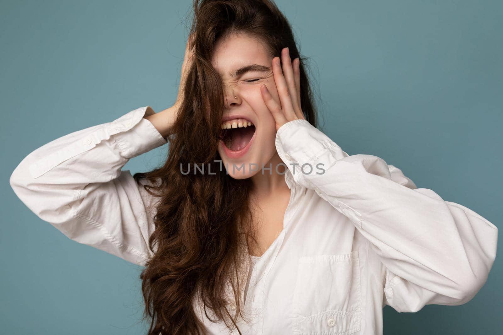 Attractive emotional cute nice adorable tender young curly brunette woman wearing white shirt isolated on blue background with copy space and shouting by TRMK
