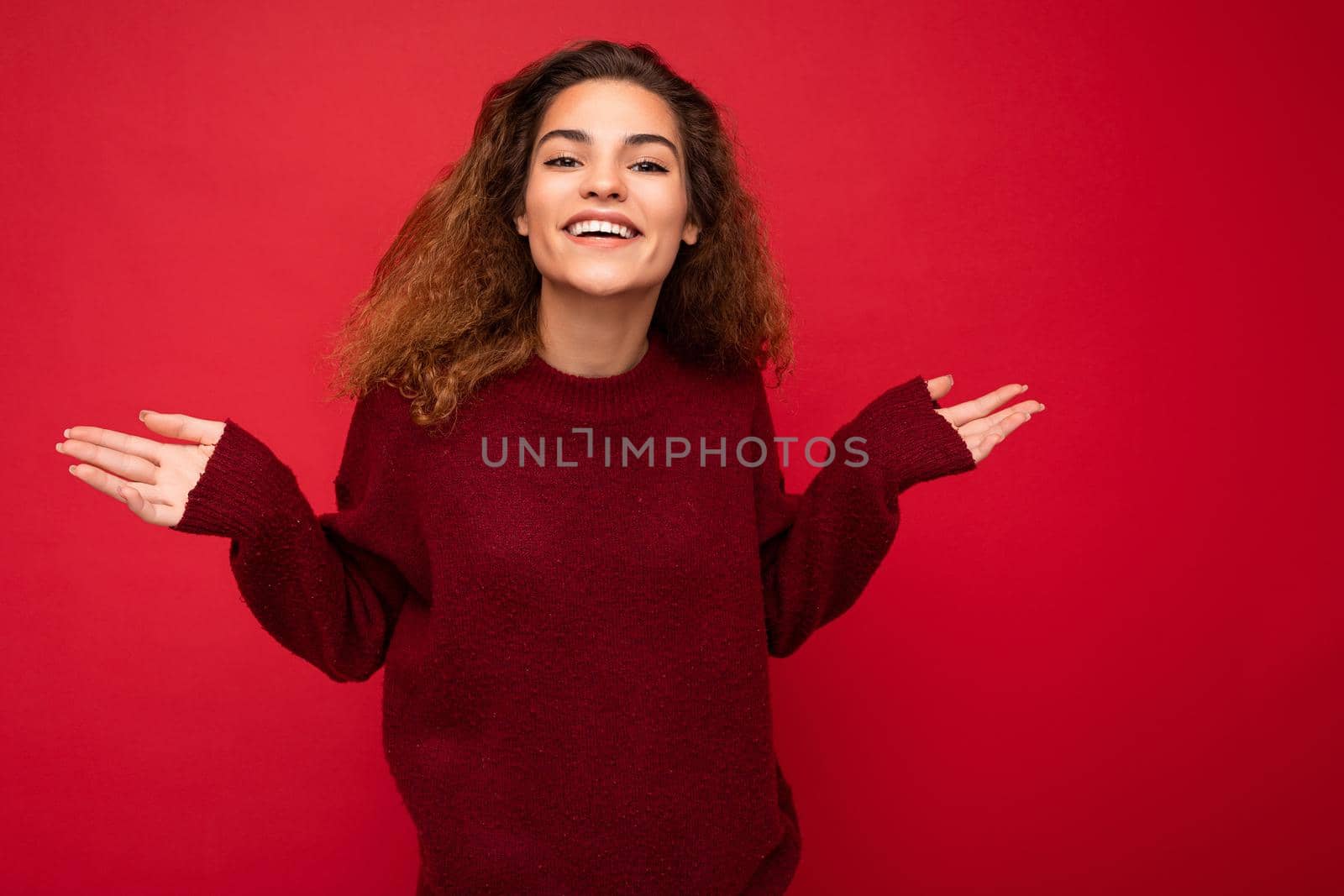 Photo of young positive happy beautiful brunette curly woman with sincere emotions wearing casual red pullover isolated on red background with copy space by TRMK