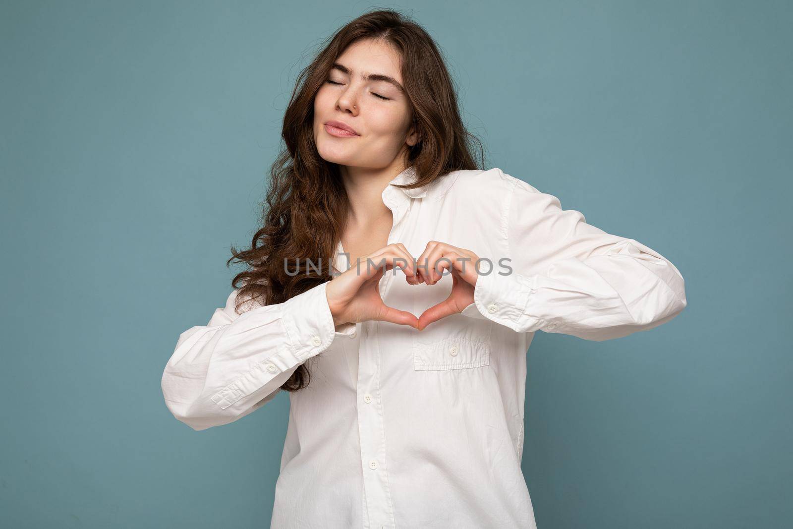 Portrait of young positive happy attractive curly brunette woman with sincere emotions wearing casual white shirt isolated on blue background with free space and making heart form with hands. Love concept.