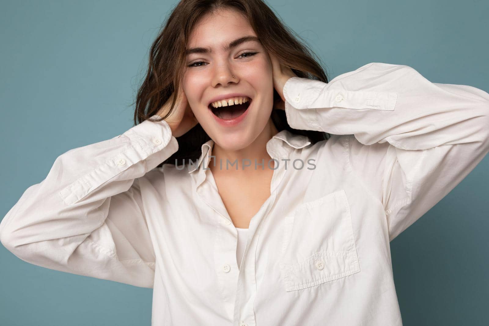 Portrait of beautiful positive cheerful cute smiling young brunette woman in casual white shirt isolated on blue background with copy space.