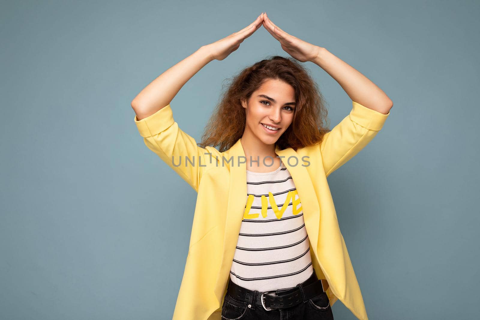 Photo shot of young positive happy attractive brunette curly woman with sincere emotions wearing trendy yellow jacket isolated on blue background with copy space and showing house gesture. Home concept.