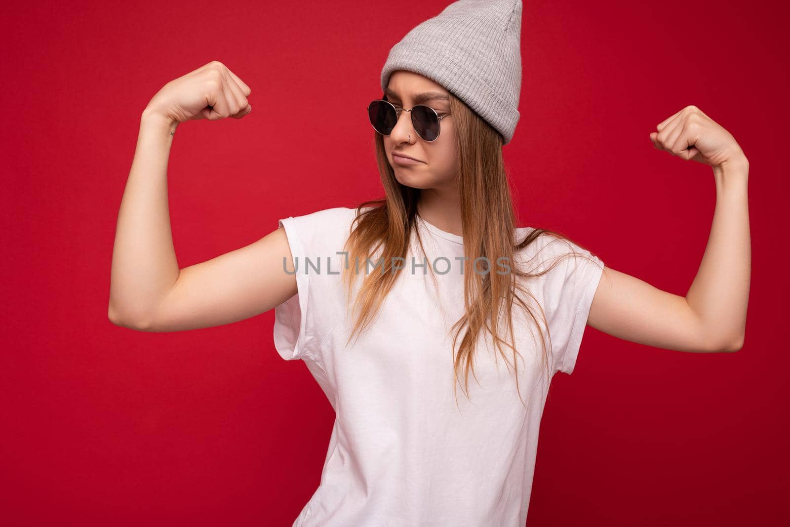 Portrait of young emotional positive funny beautiful dark blonde woman with sincere emotions wearing casual white t-shirt with empty space for mockup gray hat and sunglasses isolated over red background with free space. Female model showing powerful muscles and biceps. Fitness and workout concept.