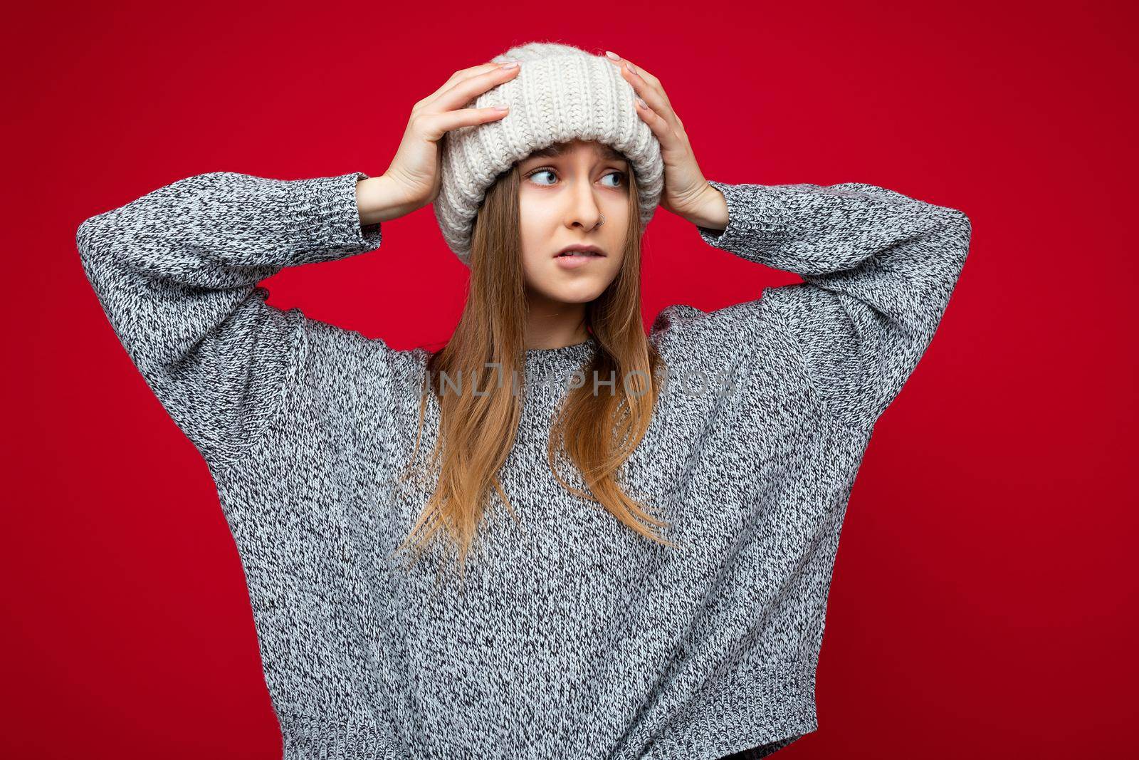 Photo of attractive upset young dark blonde female person standing isolated over red background wall wearing grey sweater and winter beige hat looking to the side and being concerned.