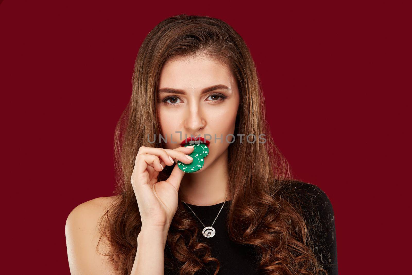 Pretty long hair woman in black dress holding chips for gambling in casino. Studio shot on red background. Poker. Two green Chips in Hand