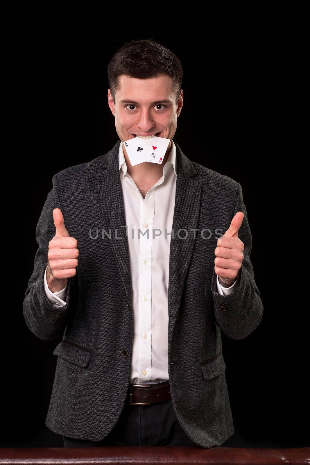 Young caucasian man in a dark suit and a white shirt holding two aces in his teeth and showing thumb up hands gesture on black background. Gambling concept. Casino