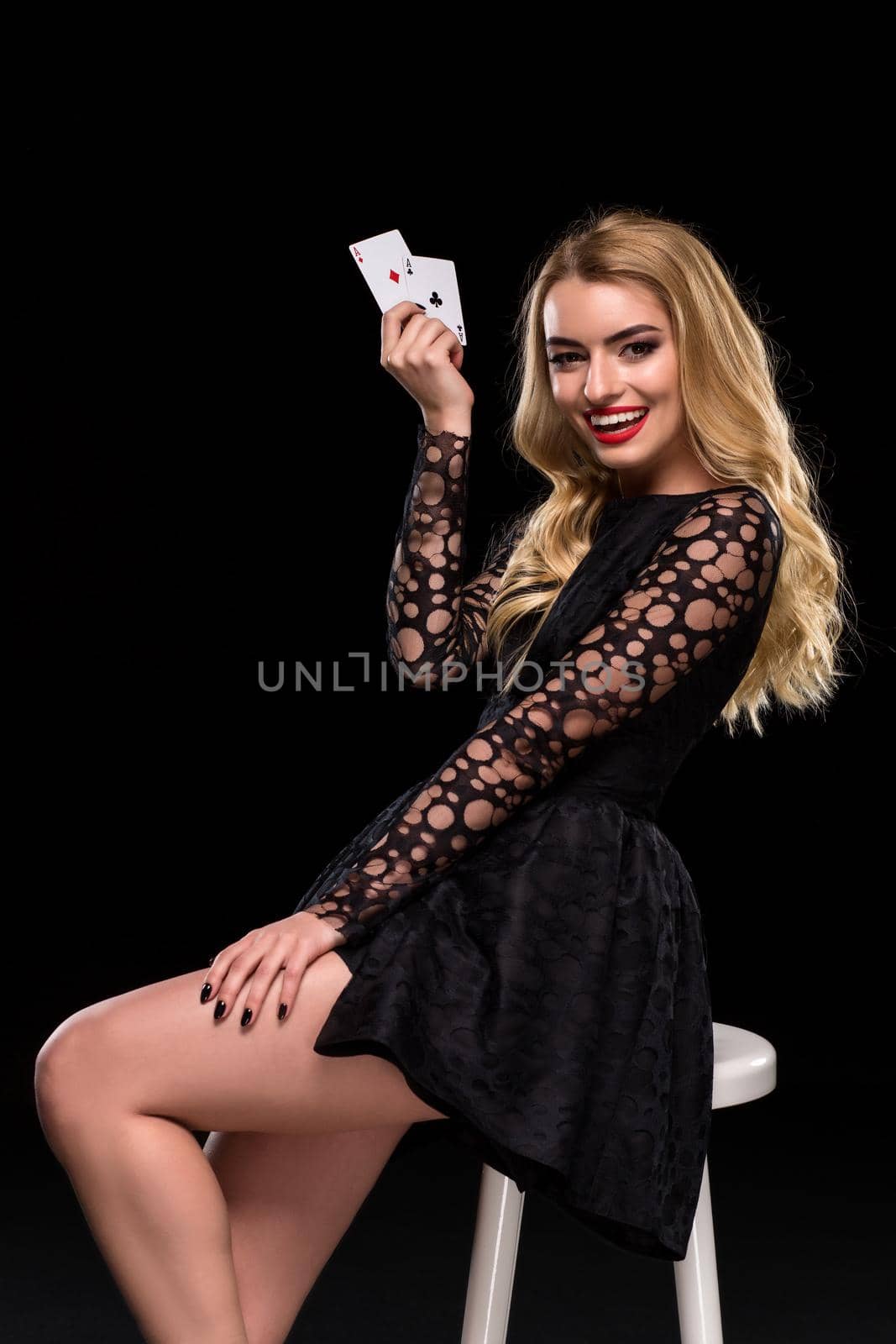 Beautiful confident woman in black dress sitting on chair showing cards looking at camera, isolated on the black background. With copy place Poker. Casino. Roulette Blackjack Spin.