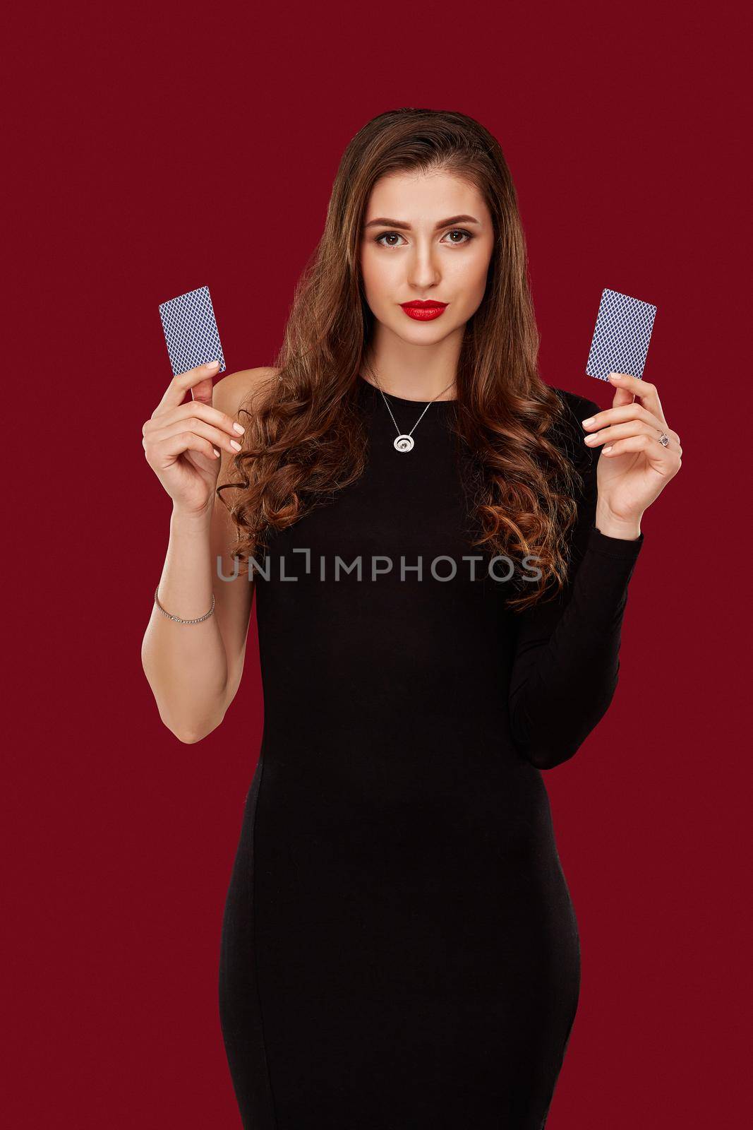 Beautiful caucasian woman in black dress with poker cards gambling in casino. Studio shot on red background. Poker. Two cards in hands