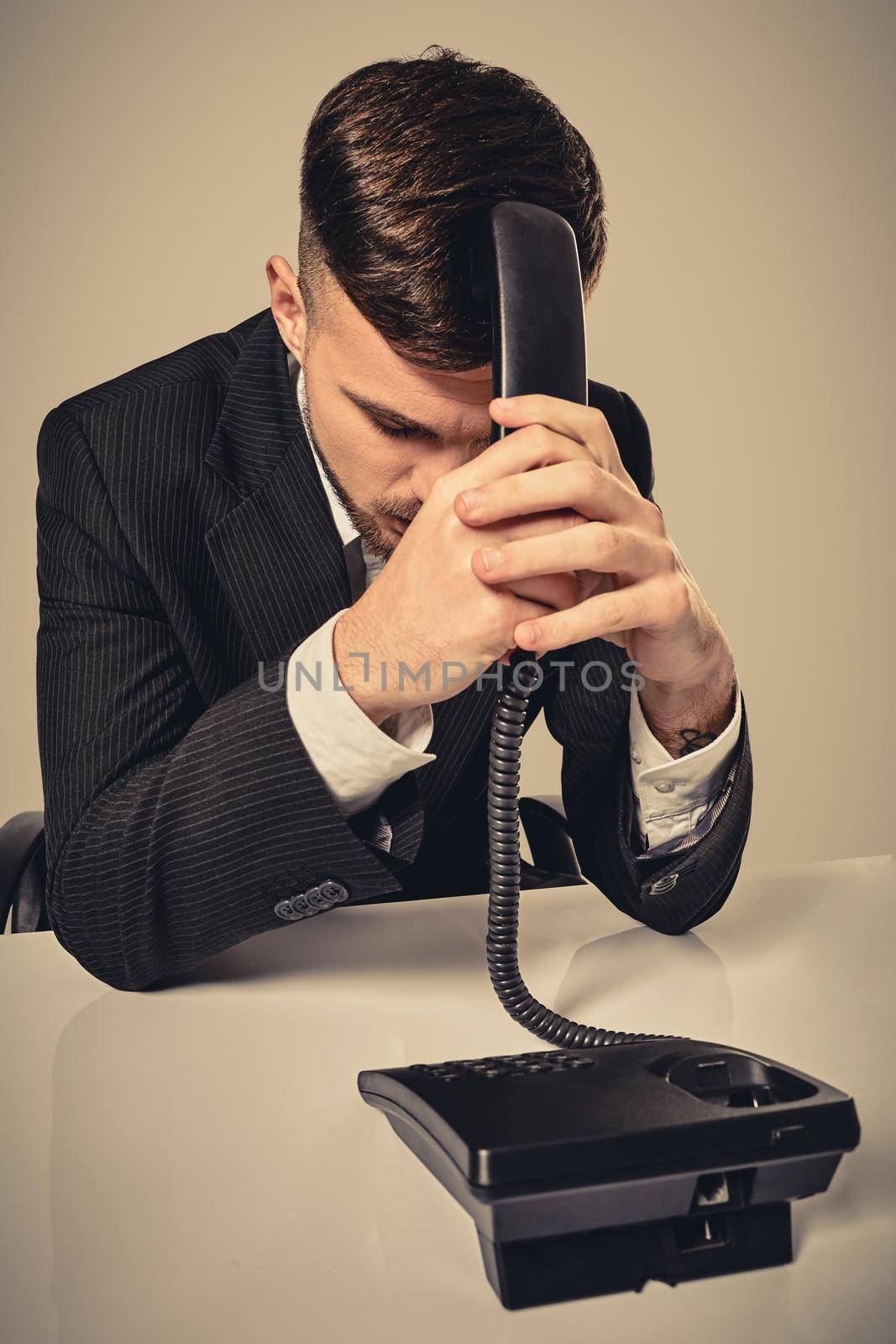 A young man in a black suit dials the phone number while sitting in the office. Manager beats himself over the head with a telephone