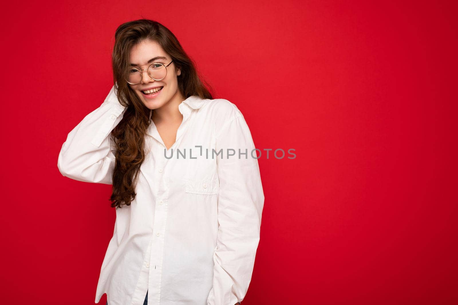 Portrait of beautiful positive cheerful cute smiling young brunette woman in casual white shirt and trendy optical glasses isolated on red background with copy space by TRMK