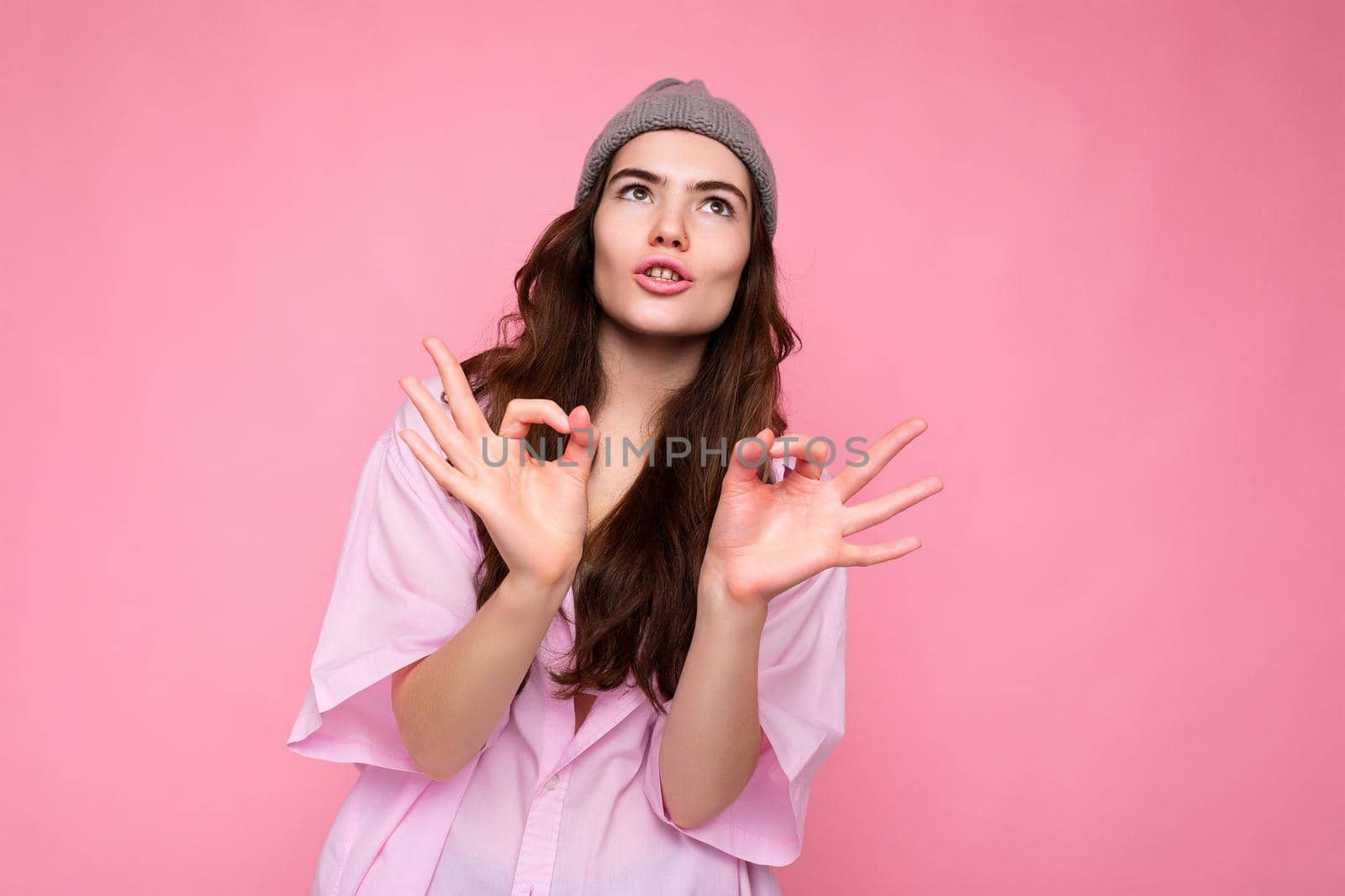 Portrait of young positive emotional funny attractive curly brunette woman with sincere emotions wearing stylish pink shirt and grey hat isolated on pink background with copy space and showing okay gesture. It's fine by TRMK