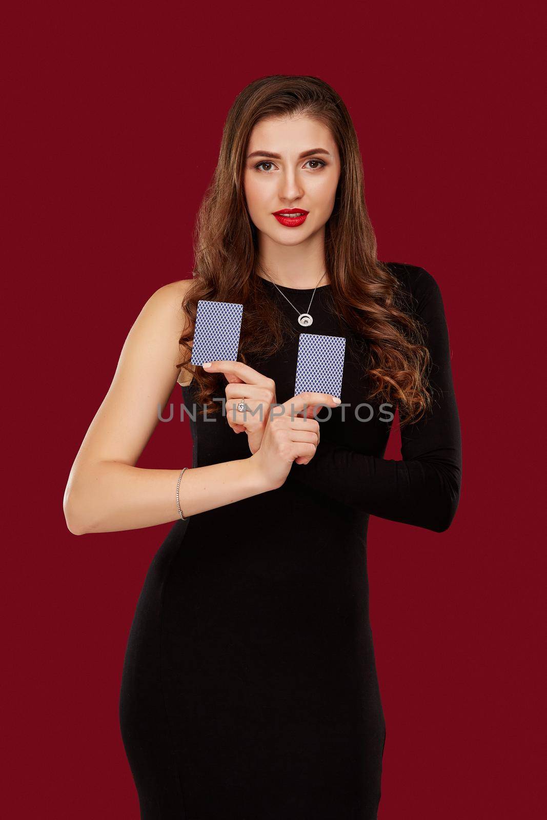 Beautiful caucasian woman in black dress with poker cards gambling in casino. Studio shot on red background. Poker. Two cards in hands