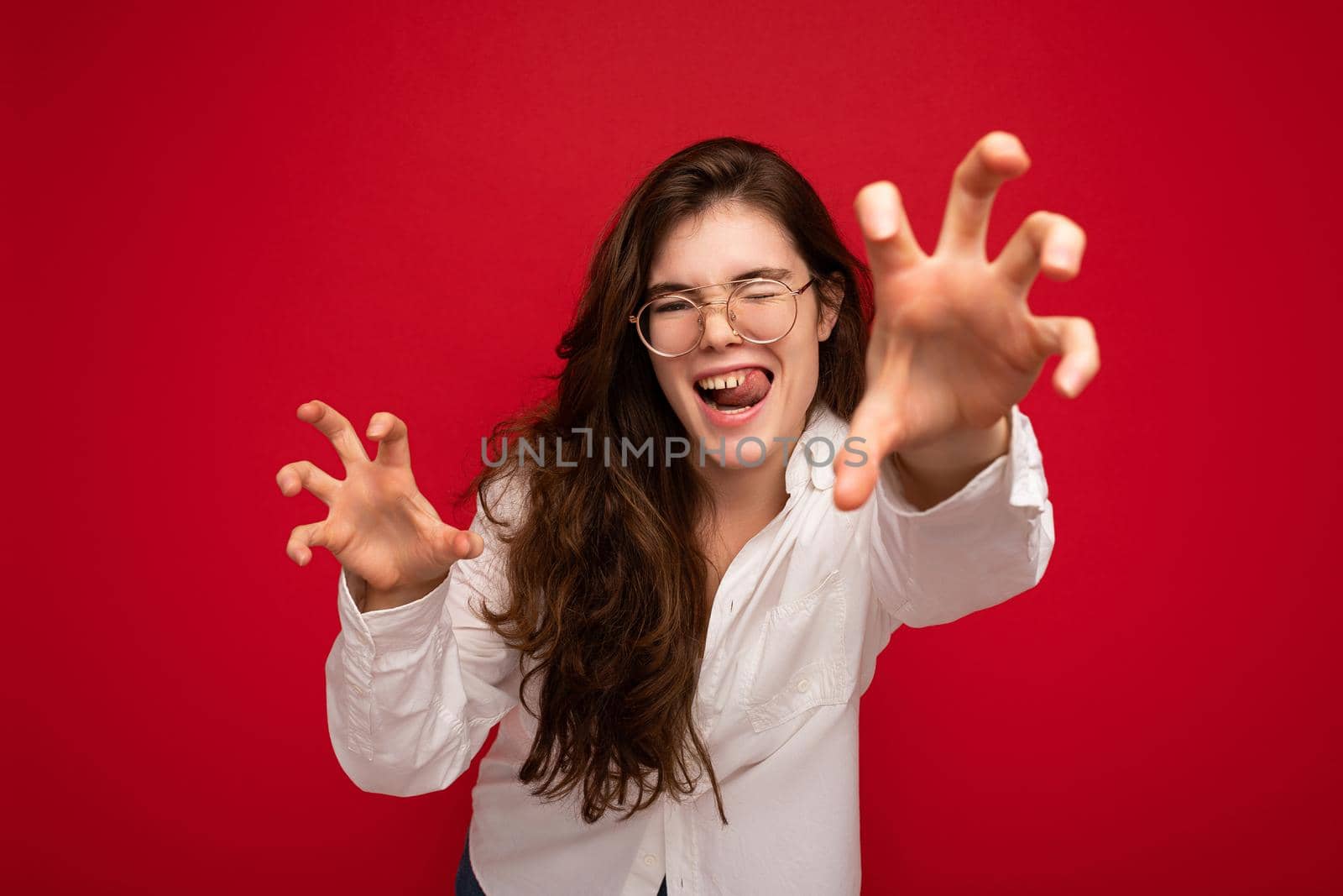 Portrait of young emotional positive sexy beautiful brunette woman with sincere emotions wearing casual white shirt and optical glasses isolated on red background with copy space and making cat claws and growling like animal by TRMK