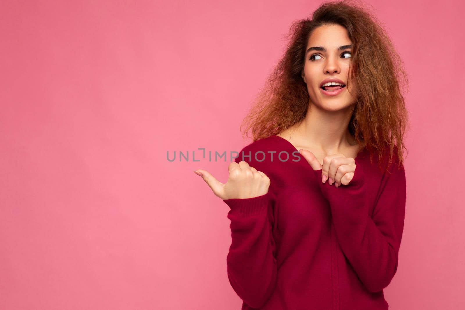 Photo of young happy positive beautiful brunette wavy woman with sincere emotions wearing casual pink jersey isolated over pink background with copy space and pointing at free space for mockup.