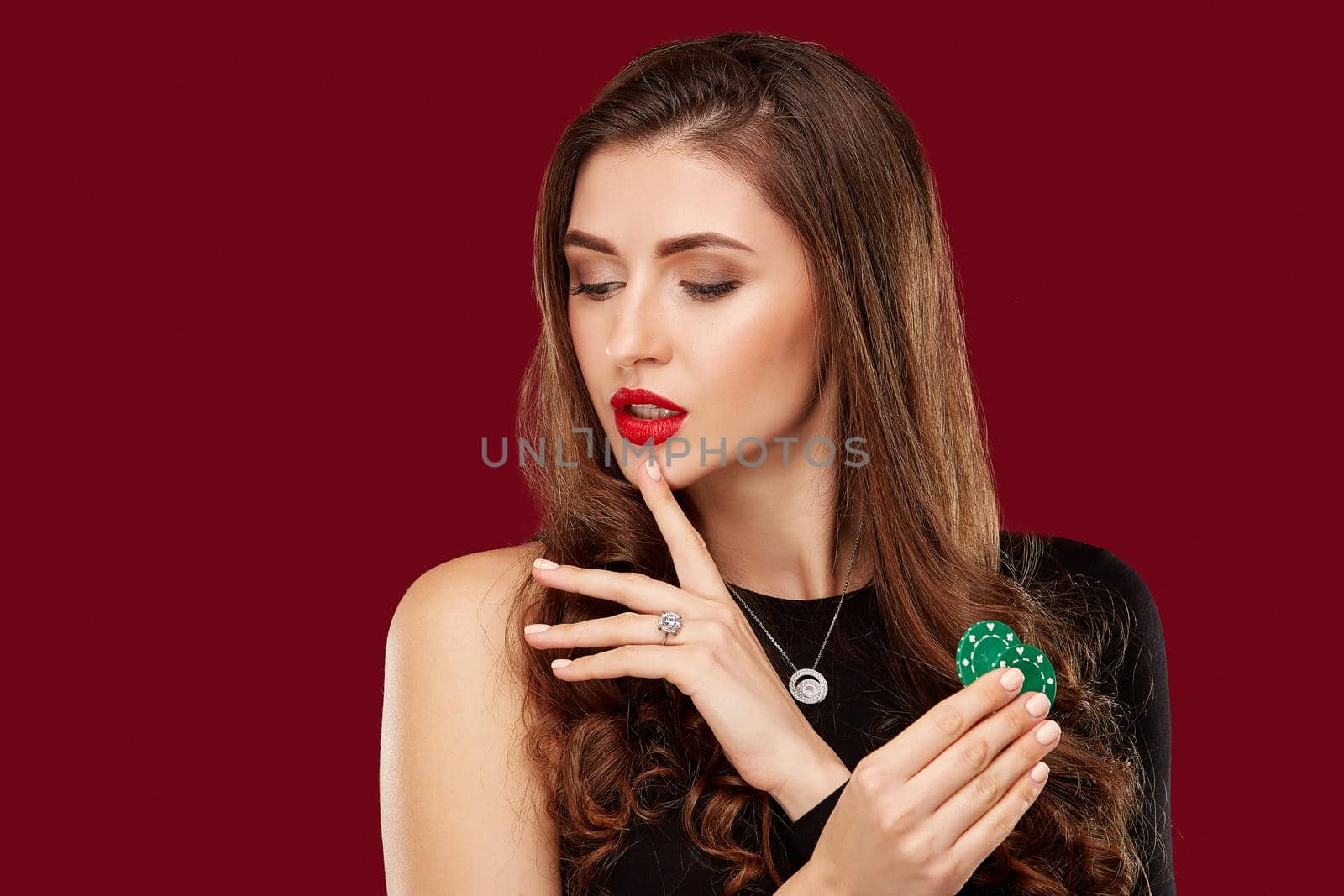 Pretty long hair woman in black dress holding chips for gambling in casino. Studio shot on red background. Poker. Two green Chips in Hand