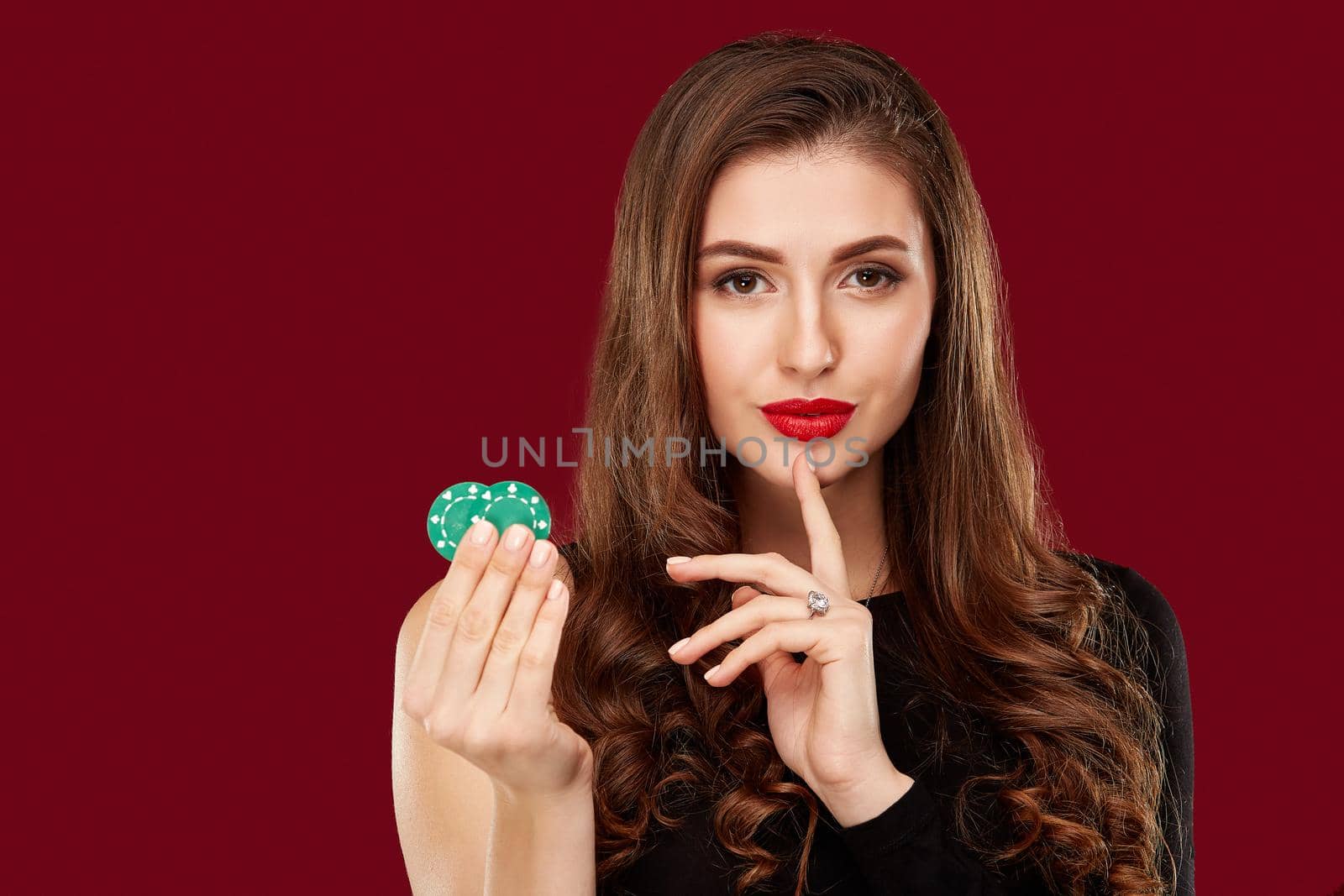 Pretty long hair woman in black dress holding chips for gambling in casino. Studio shot on red background. Poker. Two green Chips in Hand