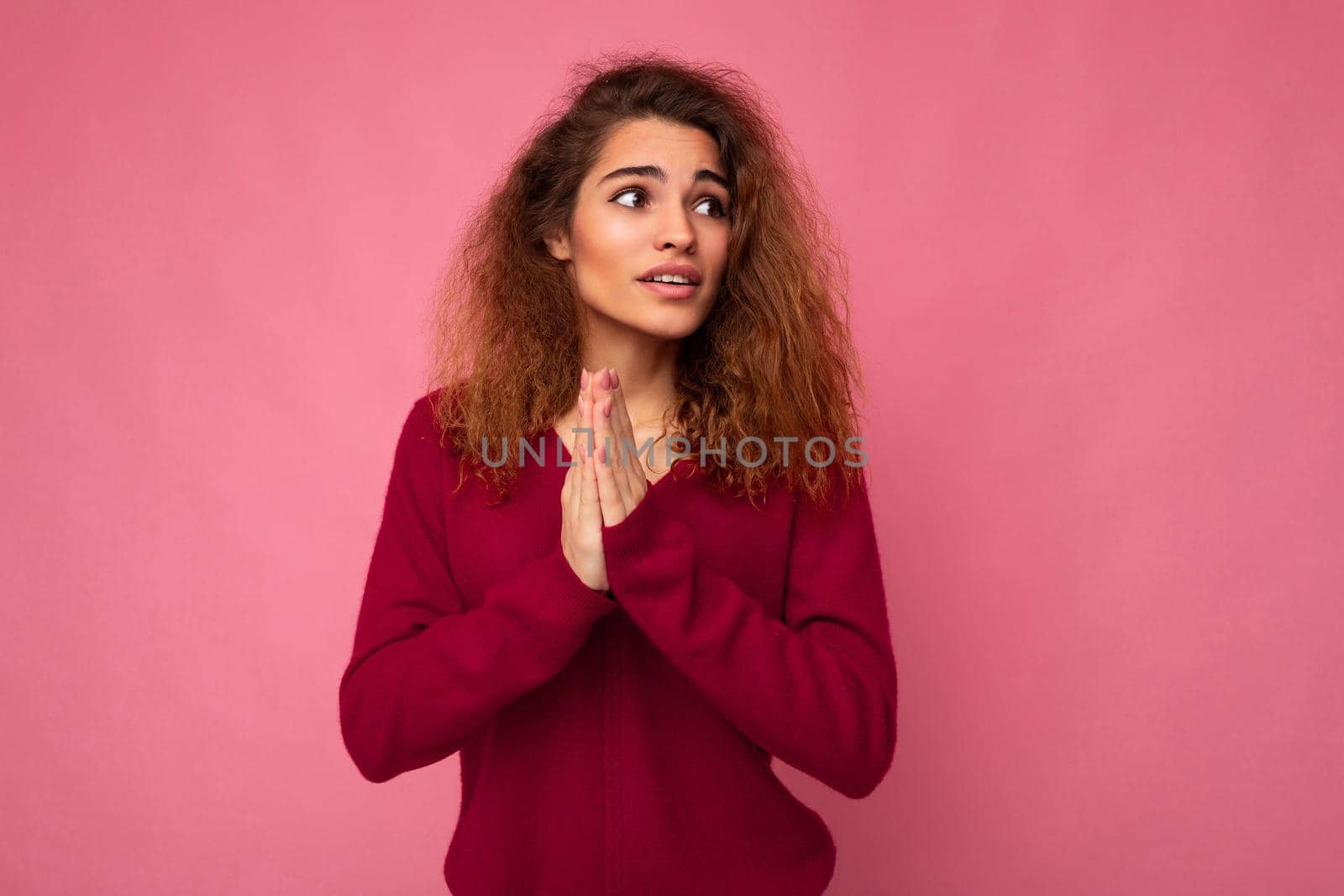 Photo of young emotional beautiful brunette curly woman with sincere emotions wearing trendy pink pullover isolated over pink background with free space and praying with hands holding together askinf for forgiveness.