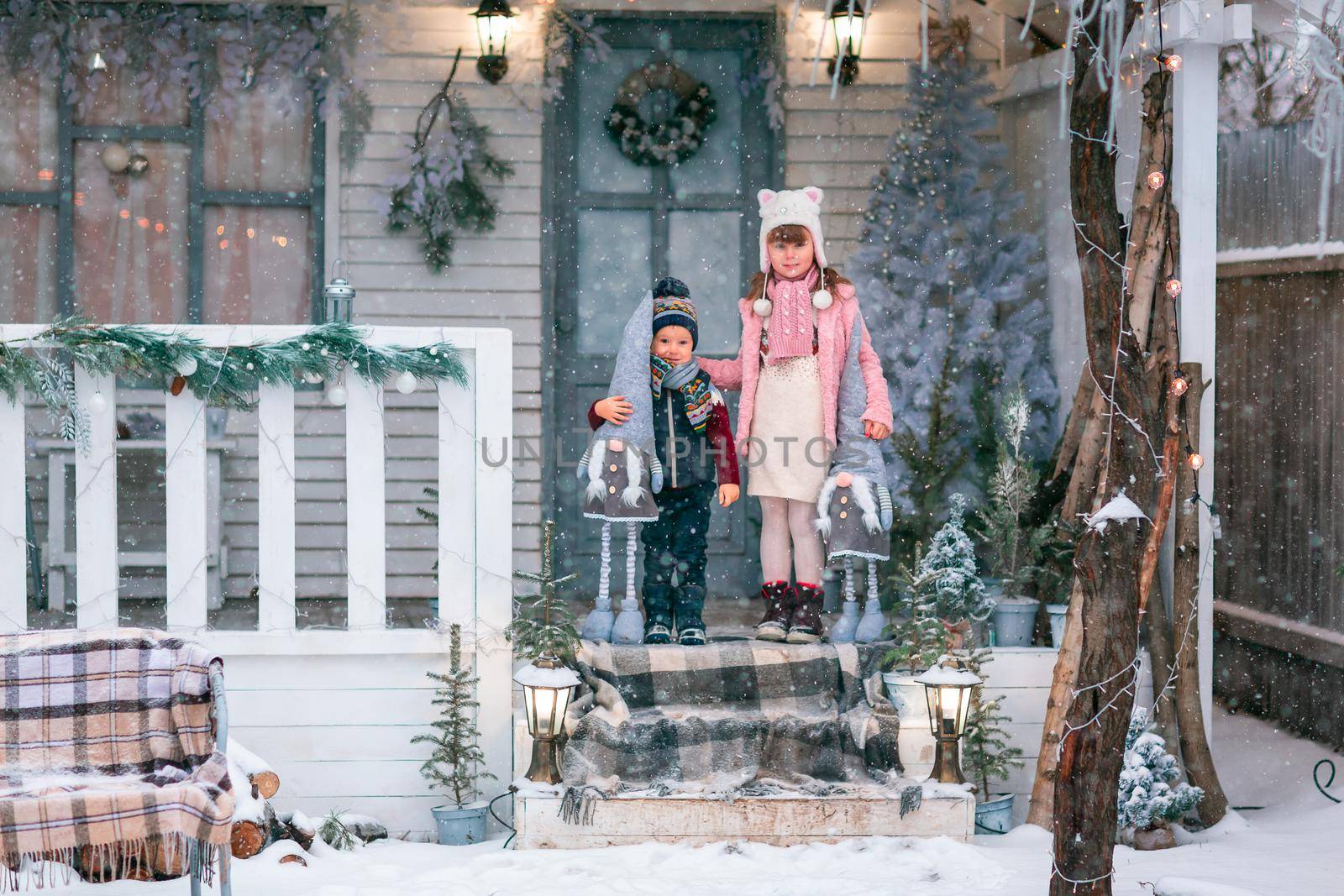 Happy little kids sitting on the porch of the Christmas decorated house, snowing outdoor. Happy New Year and Merry Christmas. Magic winter