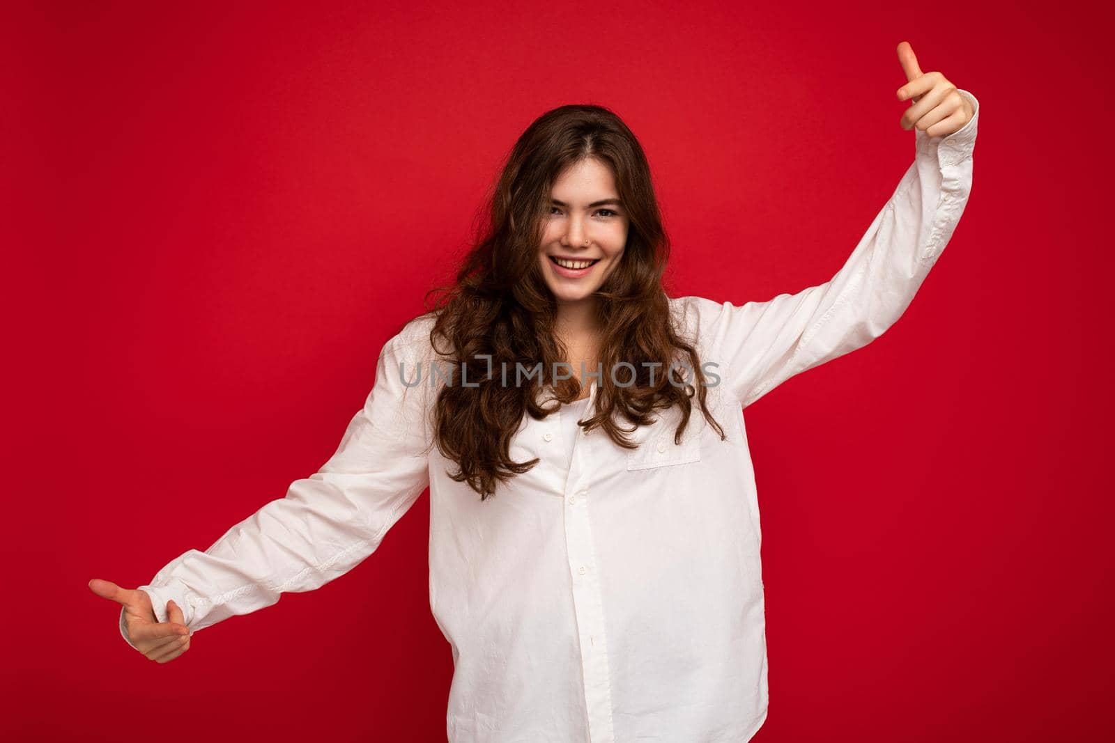 Portrait of beautiful positive cheerful cute smiling young brunette woman in casual white shirt isolated on red background with copy space.