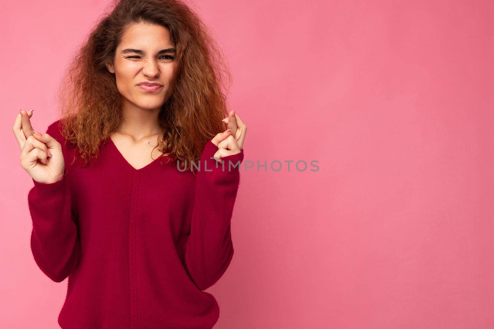 Photo shot of young attractive brunette curly woman with sincere emotions wearing trendy pink pullover isolated over pink background with empty space and keeping crossed fingers. Gesture concept.