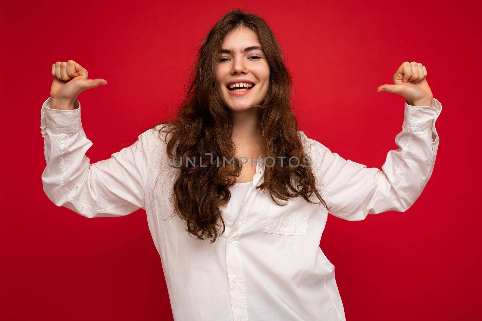 Photo of young beautiful happy smiling curly brunette woman wearing white shirt. Sexy carefree female person posing isolated near red wall in studio with free space. Positive model with natural makeup.