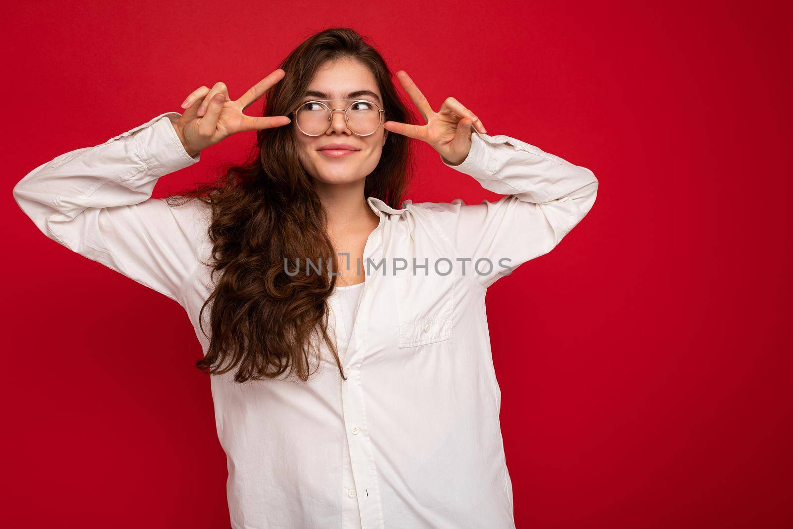 Photo of young beautiful happy smiling brunette woman wearing white shirt and optical glasses. Sexy carefree female person posing isolated near red wall in studio with free space. Positive model with natural makeup.
