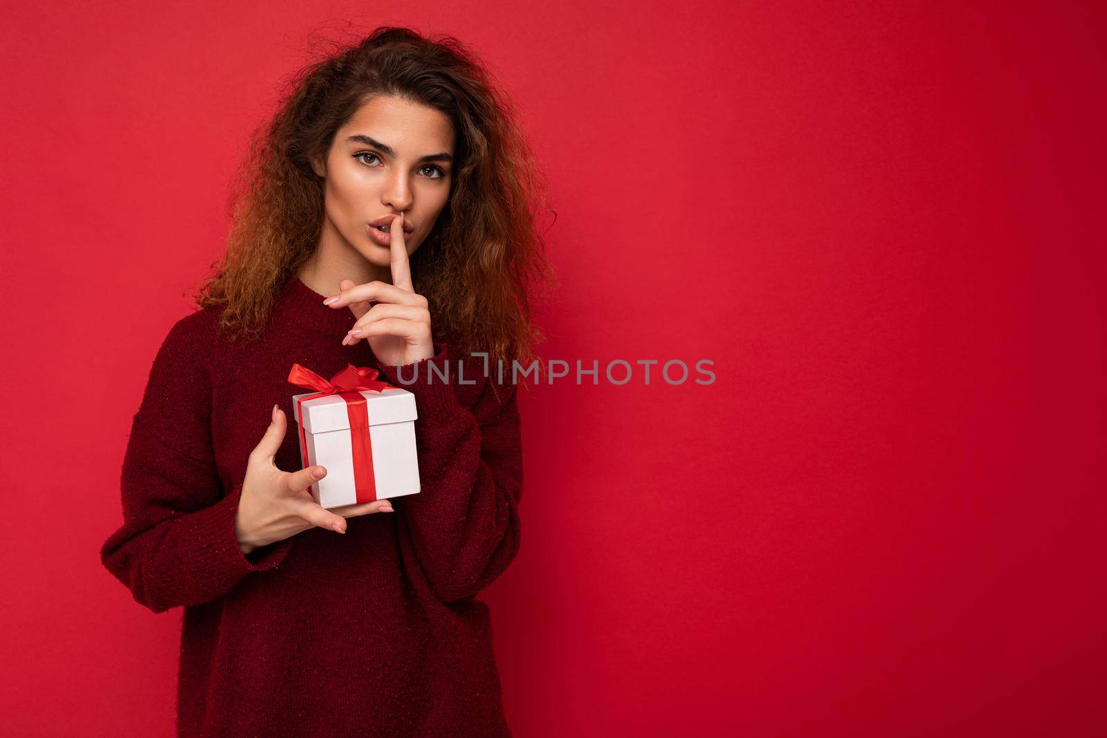 Shot of pretty young brunette curly woman isolated over red background wall wearing red sweater holding gift box looking at camera and showing shh gesture and meaning be quiet. Copy space, mockup