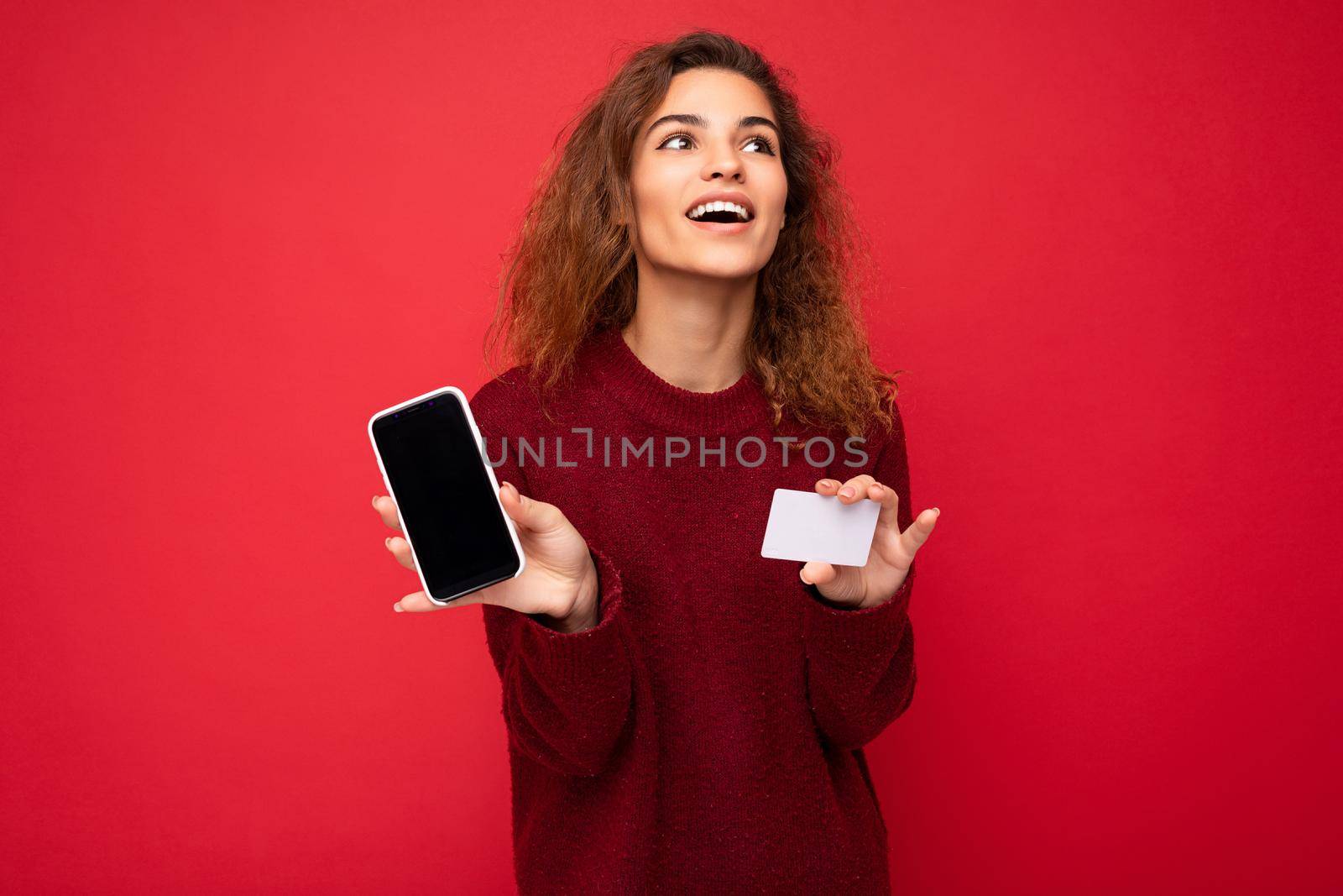 Young beautiful curly brunette woman with sincere emotions poising isolated over background wall with empty space wearing casual dark red sweater holding credit card mobile phone with empty screen display for mock up. Internet concept.