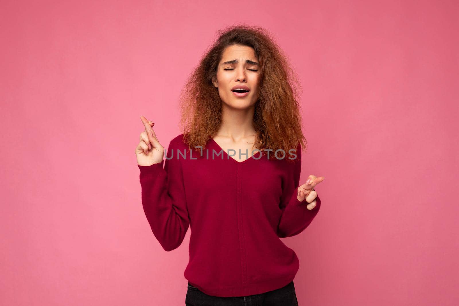 Photo shot of young attractive brunette curly woman with sincere emotions wearing trendy pink sweater isolated over pink background with empty space and holding fingers crossed for good luck. Gesture concept.
