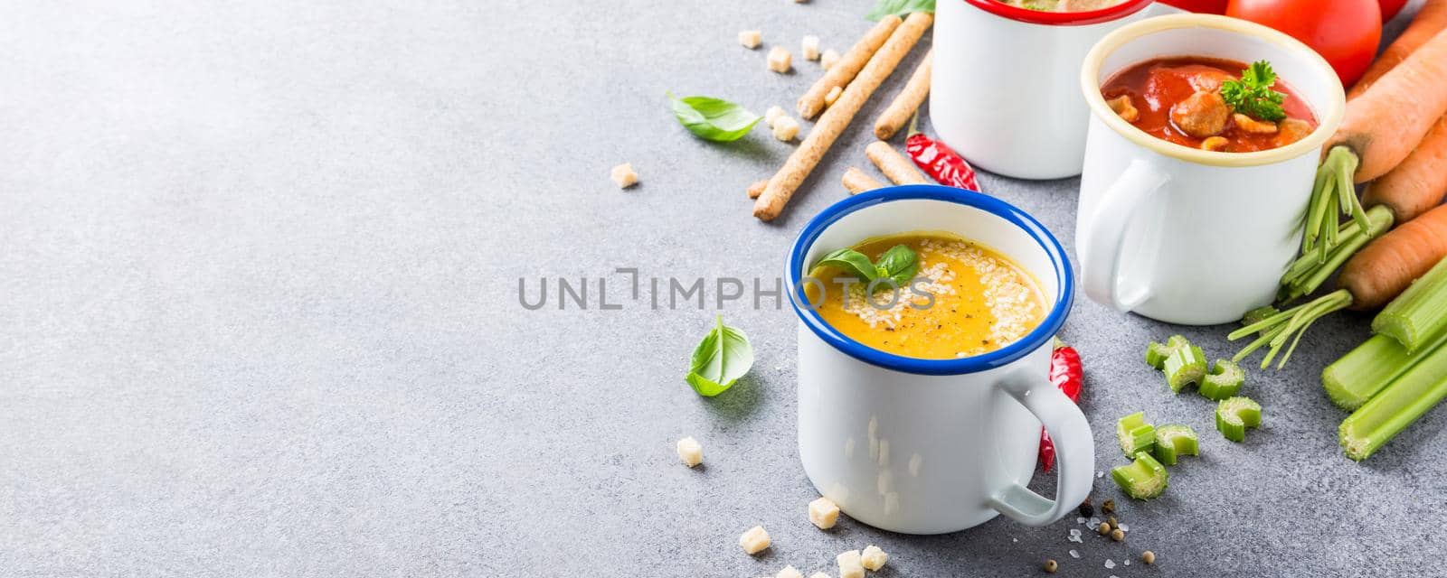 Assorted delicious homemade soups in enamel mugs with ingredients. Healthy food concept with copy space.