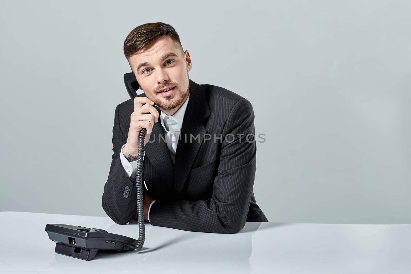 A young man in a black suit dials the phone number while sitting in the office. Manager looking at the camera and talking on the phone