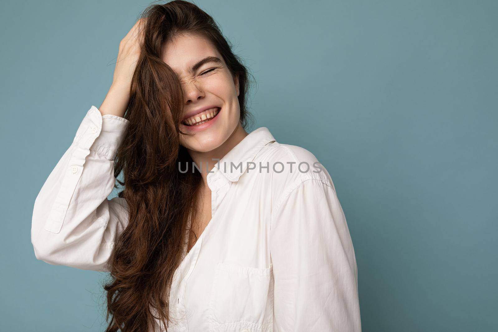 Portrait of beautiful positive cheerful cute smiling young brunette woman in casual white shirt isolated on blue background with copy space by TRMK
