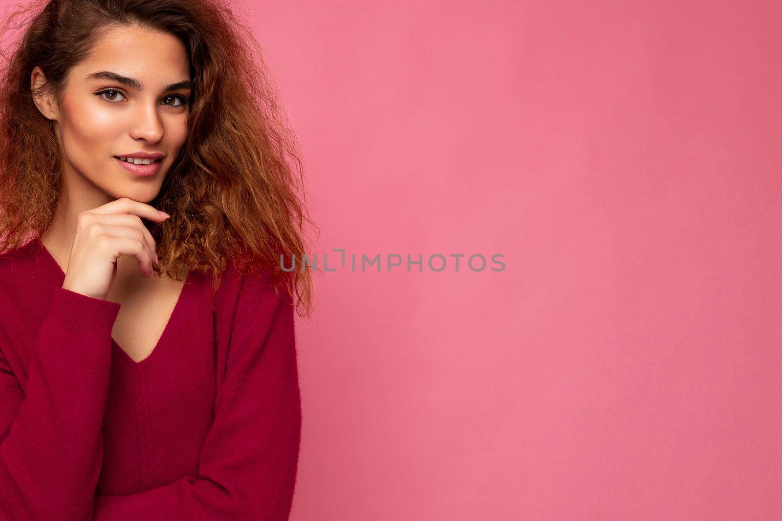 Portrait of young positive thoughtful beautiful brunette curly woman with sincere emotions wearing trendy pink pullover isolated on pink background with copy space.