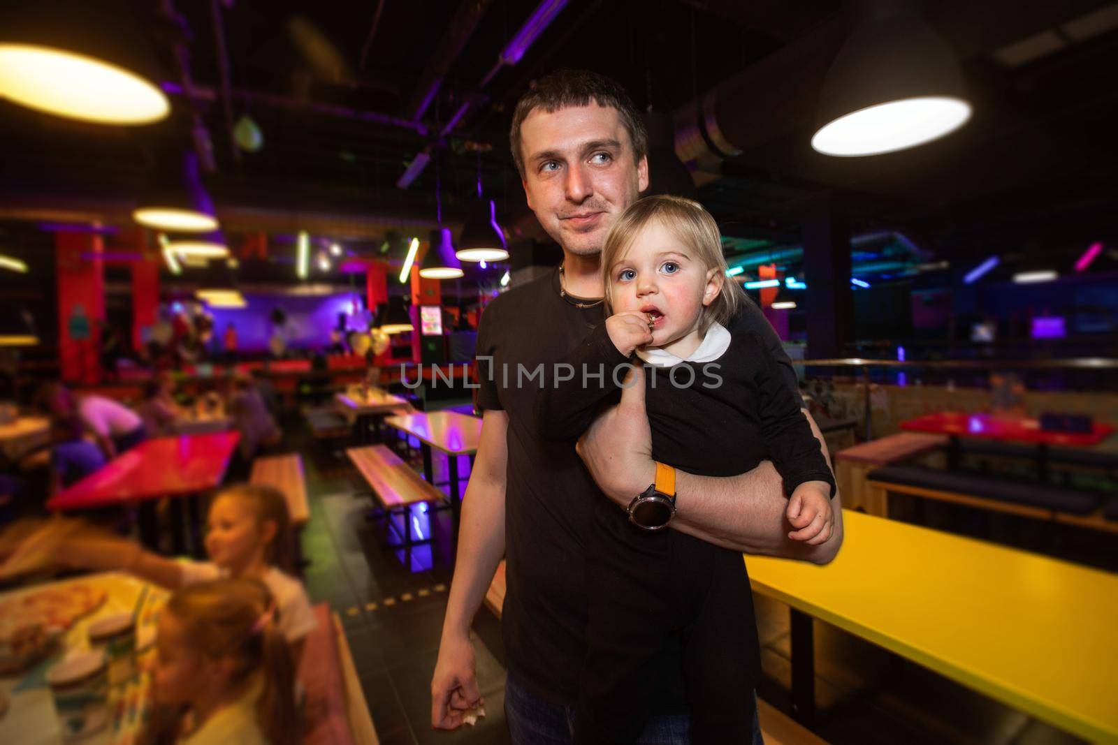 Adult man carrying cute girl and looking away while standing in illuminated cafe of family entertainment center during children party