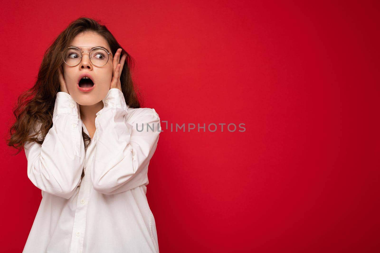 Attractive shocked amazed surprised young curly brunette woman wearing white shirt and optical glasses isolated on red background with empty space.